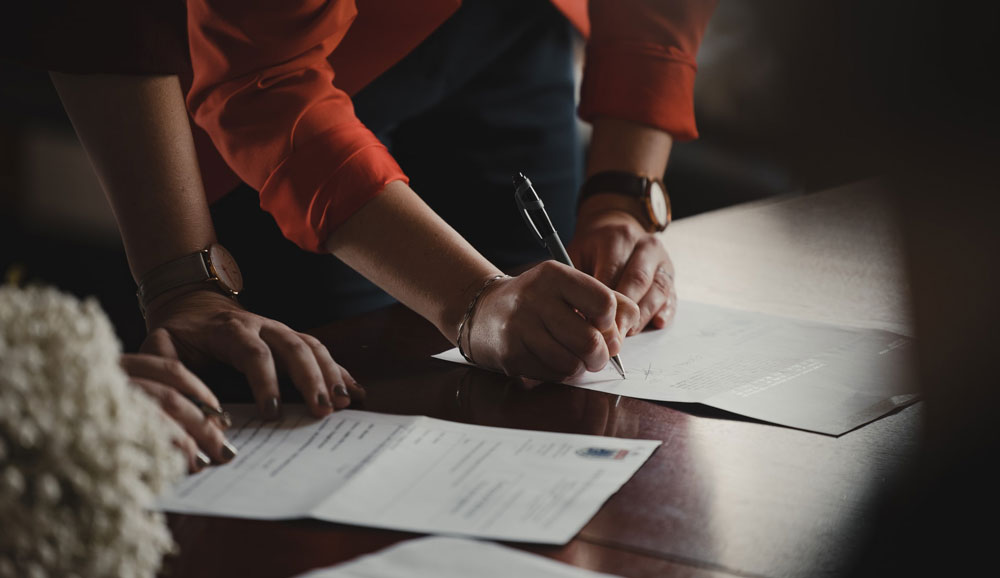 two-people-filling-out-paperwork Close up of two people's arms as they lean over a desk to sign paperwork